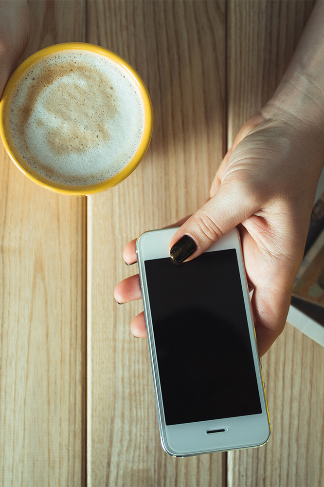 Hand holding a smartphone and a cup of coffee on a wooden table, representing a casual moment for someone checking real estate opportunities. We buy northeast Ohio houses in any condition.